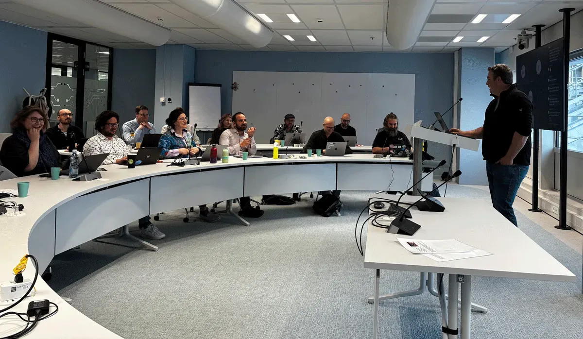 Meeting attendees seated around a large curved table with laptops, listening to a person speaking at a lectern
